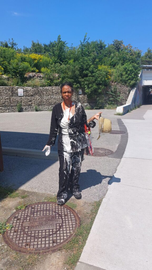 A Black woman, dressed in all black clothing, carrying a sun hat, covered in white paint.